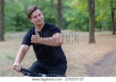 Young Handsome Caucasian Man Thumbs Up Bicycling At The Park. White Earphones, Dark Casual Wear. Out