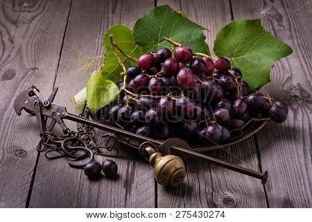 Still Life With Black Grapes In An Old Steelyard On Rough Wooden Table