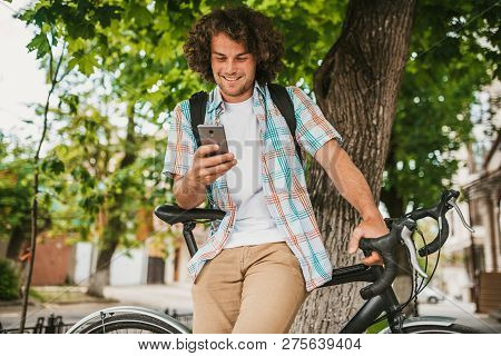 Happy Young Male With Curly Hair Wearing Shirt With Backpack, Sitting On The Bike On The City Street