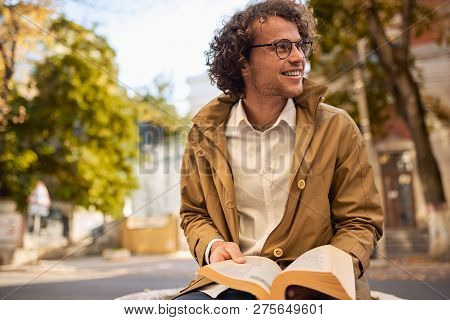 Happy Young Man With Glasses Reading And Posing With Book Outdoors. College Male Student Carrying Bo