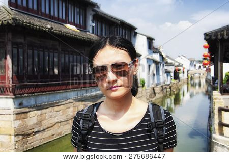 A Chinese Woman Wearing Sunglasses On A Bridge With The Buildings Of Luzhi Water Town Blurred In The