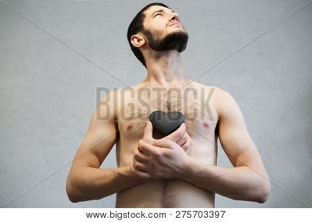 Naked Young, Bearded Man, Looking Up And Holding Black Heart In Hands Near Torso.