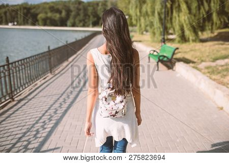 Beautiful Back View Of Young Bruntte Woman With Long Hair And With Backpack, Walking In The Park Wit