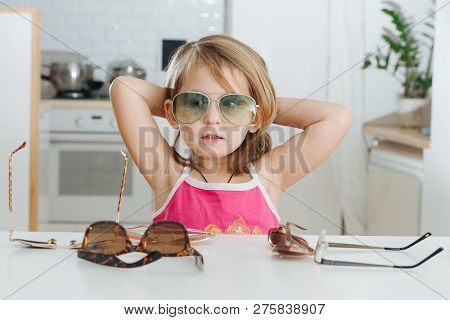 Portrait Of Cute Dark Blonde Little Girl With Short Hair Trying To Wear Blue Aviator Sunglasses. She