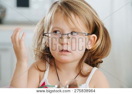 Portrait Of Cute Dark Blonde Little Girl With Short Hair Trying To Wear Pinhole Glasses. She Is At H