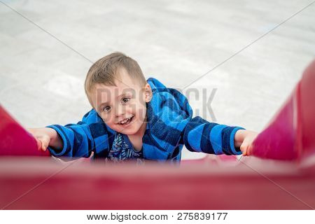 Little Cheeky Mischevious Caucasian Boy Climbing Up The Red Plastic Outdoor Slide On A Playground