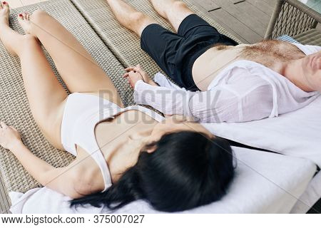 Young Couple Enjoying Resting On Chaise-lounges By Swimming Pool, Thay Are Holding Hands And Looking