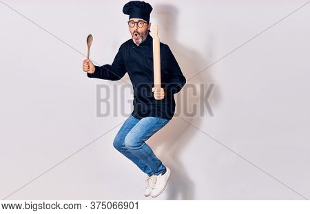 Middle age handsome hispanic man surprised wearing glasses and cook uniform. Holding kneader and spoon jumping over isolated white background