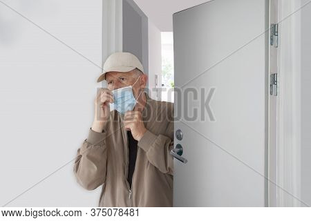 Man With Cap Ajusting His Protective Medical Mask As He Leaves Home. He Walks Out A Half Opened Door