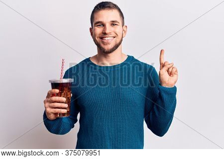 Young handsome man dirnking cola refreshment beverage smiling with an idea or question pointing finger with happy face, number one 