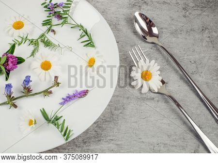 Flowers And Green Plant In A White Plate With Fork And Spoon On Grey Textured Background, Vegetarian