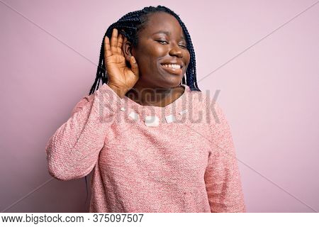 African american plus size woman with braids wearing casual sweater over pink background smiling with hand over ear listening an hearing to rumor or gossip. Deafness concept.
