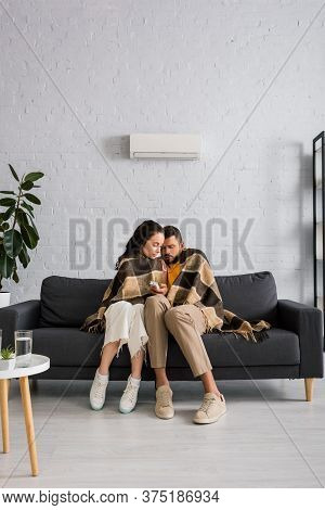 Wrapping In Plaid And Holding Remote Controller Of Air Conditioner While Feeling Cold On Sofa