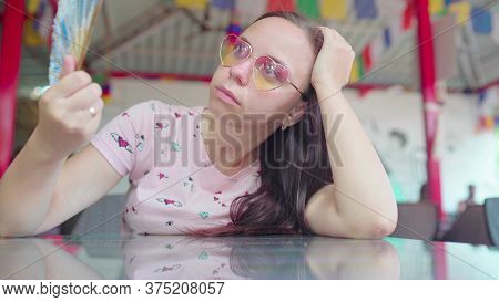 A Beautiful Young Woman Is Waving A Fan In The Sweltering Heat In A Tropical Cafe.