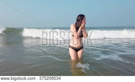 A Young Woman In A Black Swimsuit Spends Time In The Sea Or Ocean. The Girl Takes Refuge From The Wa