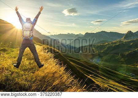 Hiking Man With Backpack Jumping With Raised Arms On Mountain. Beautiful Sunrise And Layered Mountai