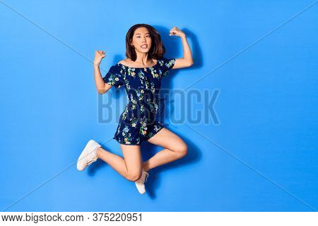 Young beautiful chinese woman wearing casual clothes smiling happy. Jumping with smile on face doing strong geture over isolated blue background