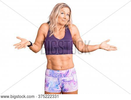 Middle age fit blonde woman wearing sportswear clueless and confused expression with arms and hands raised. doubt concept. 