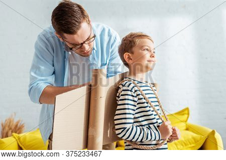 Father Fixing Carton Plane Wings On Back Of Happy Son