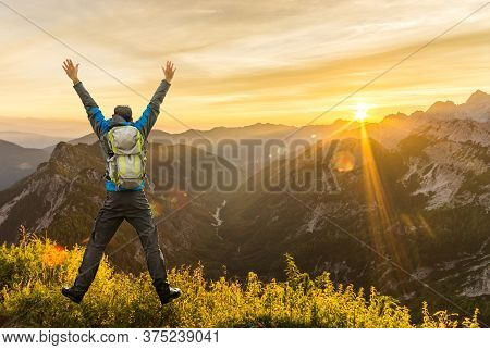 Hiking Man With Backpack Jumping With Raised Arms On Mountain. Amazing Sunrise Backlight With Beauti