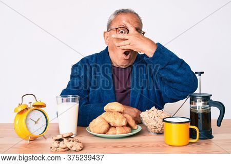 Senior handsome man with gray hair sitting on the table eating breakfast in the morning peeking in shock covering face and eyes with hand, looking through fingers afraid 