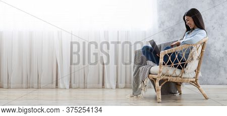 Freelance Concept. Asian Girl Sitting In Wicker Chair With Laptop Working Remotely At Home, Panorama