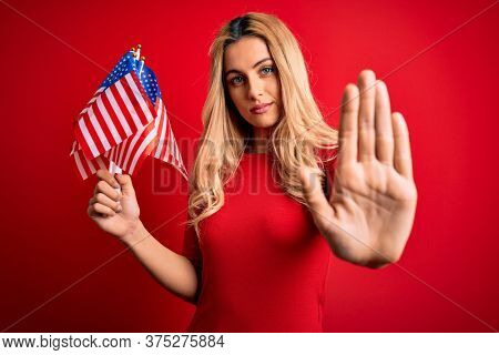 Beautiful blonde patriotic woman holding united states flags celebrating independence day with open hand doing stop sign with serious and confident expression, defense gesture