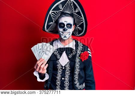 Young man wearing mexican day of the dead costume holding dollars looking positive and happy standing and smiling with a confident smile showing teeth 