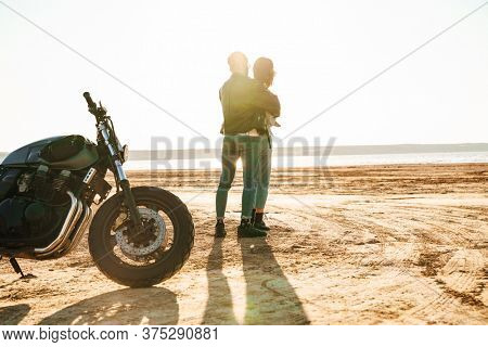 Beautiful young cople wearing jackets spending time at the sunny beach, standing near a motorbike