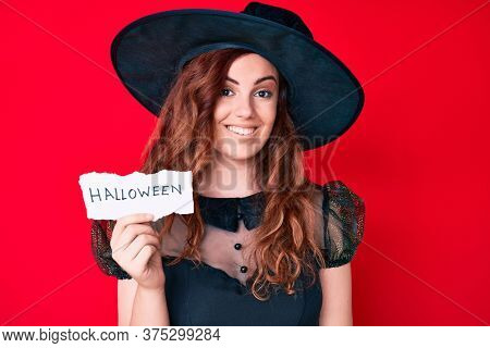 Young beautiful woman wearing witch halloween costume holding paper with halloween message looking positive and happy standing and smiling with a confident smile showing teeth 