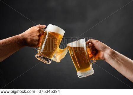 Two Hands With Beer Mugs, Toasting In Celebration, With Overflowing Foam, Dark Background And Space