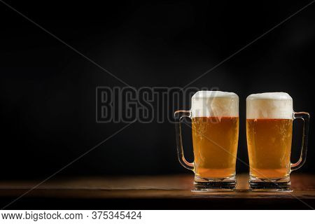 Two Cold Mugs With Beer, With Overflowing Foam, On Wooden Table And Dark Background, Space For Writi