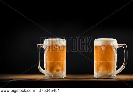 Two Cold Mugs With Beer, With Overflowing Foam, On Wooden Table And Dark Background, Space For Writi