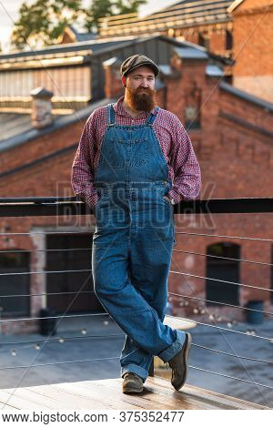 Portrait Of Brutal Bearded Motorcycle Repairman Wearing Blue Overalls, Checked Shirt And Cap In Vint