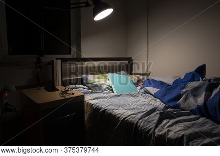 Untidy Single Bedroom. The Desk Lamp Is On. The Photo Was Taken At Night.