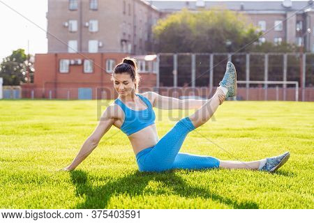 Beautiful, Athletic Caucasian Girl Sitting On A Twine In A Green Grassy Sports Stadium. Making Sport
