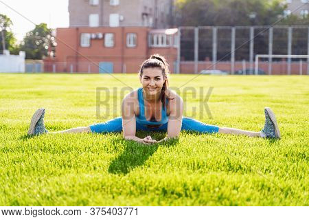 Beautiful, Athletic Caucasian Girl Sitting On A Twine In A Green Grassy Sports Stadium. Making Sport