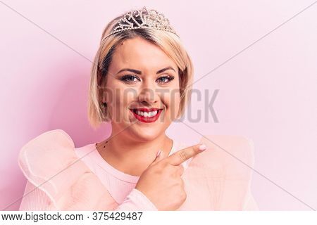 Beautiful blonde plus size woman wearing pincess tiara with diamonds over pink background smiling cheerful pointing with hand and finger up to the side