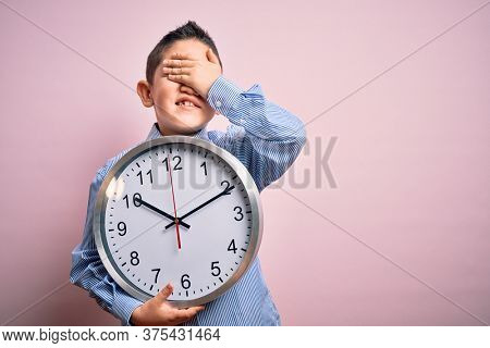 Young little boy kid holding big minute clock over isolated pink background stressed with hand on head, shocked with shame and surprise face, angry and frustrated. Fear and upset for mistake.