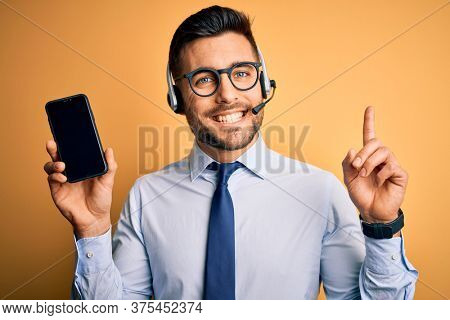 Business operator man with customer service headset from call center showing smartphone screen surprised with an idea or question pointing finger with happy face, number one
