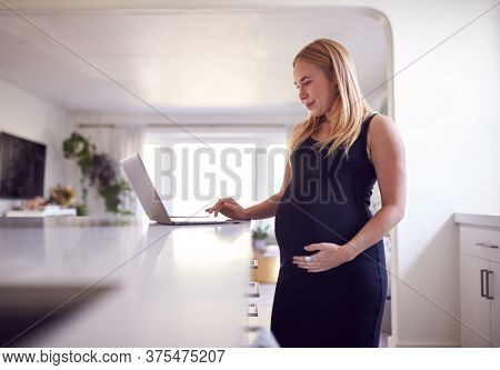Pregnant Woman Standing By Kitchen Counter Working From Home On Laptop