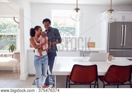 Family With Baby Daughter In Kitchen Using Laptop On Counter