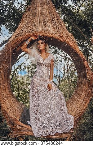 Beautiful Bride In Her Wedding Dress Posing For Photos On A Wooden Swing In The Tree.