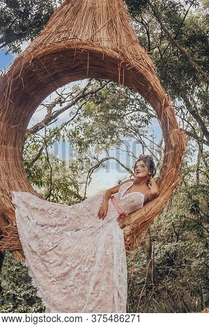 Beautiful Bride In Her Wedding Dress Posing For Photos On A Wooden Swing In The Tree.