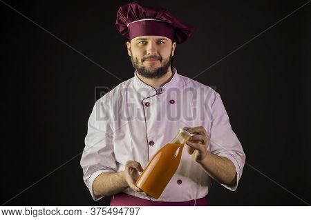 Smiling Young Bearded Male Chef Cook In Uniform Holding Plastic Bottle Of Fruit Juice Looking At Cam