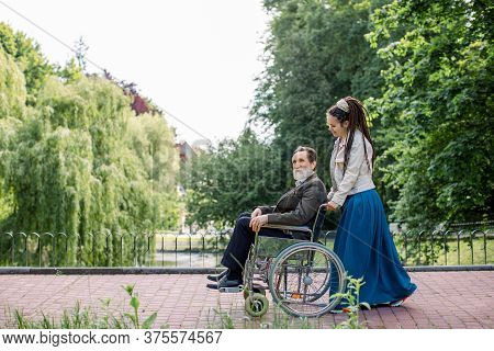 Side View Of Young Modern Hipster Woman With Long Dreadlocks, Assisting Her Disabled Grandfather On 