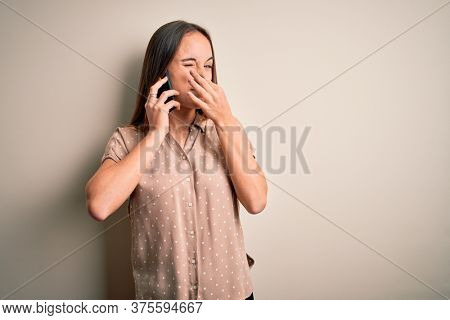 Young beautiful woman having conversation talking on the smartphone over white background smelling something stinky and disgusting, intolerable smell, holding breath with fingers on nose. Bad smell