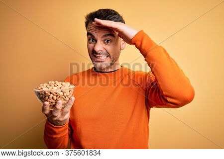 Young handsome man holding bowl with peanuts standing over isolated yellow background stressed with hand on head, shocked with shame and surprise face, angry and frustrated. Fear and upset for mistake