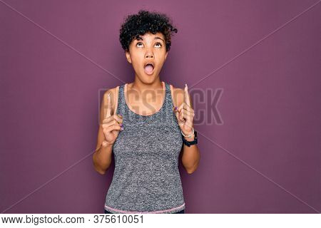 Young beautiful african american afro sportswoman doing exercise wearing sportswear amazed and surprised looking up and pointing with fingers and raised arms.