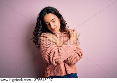 Young beautiful woman with curly hair wearing casual sweater over isolated pink background Hugging oneself happy and positive, smiling confident. Self love and self care